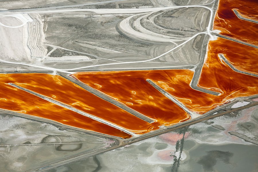 Aerial photo of the mineral pools of Searles Lake shimmering with fiery reds and oranges amidst a vast expanse of muted beige terrain, near Trona, California