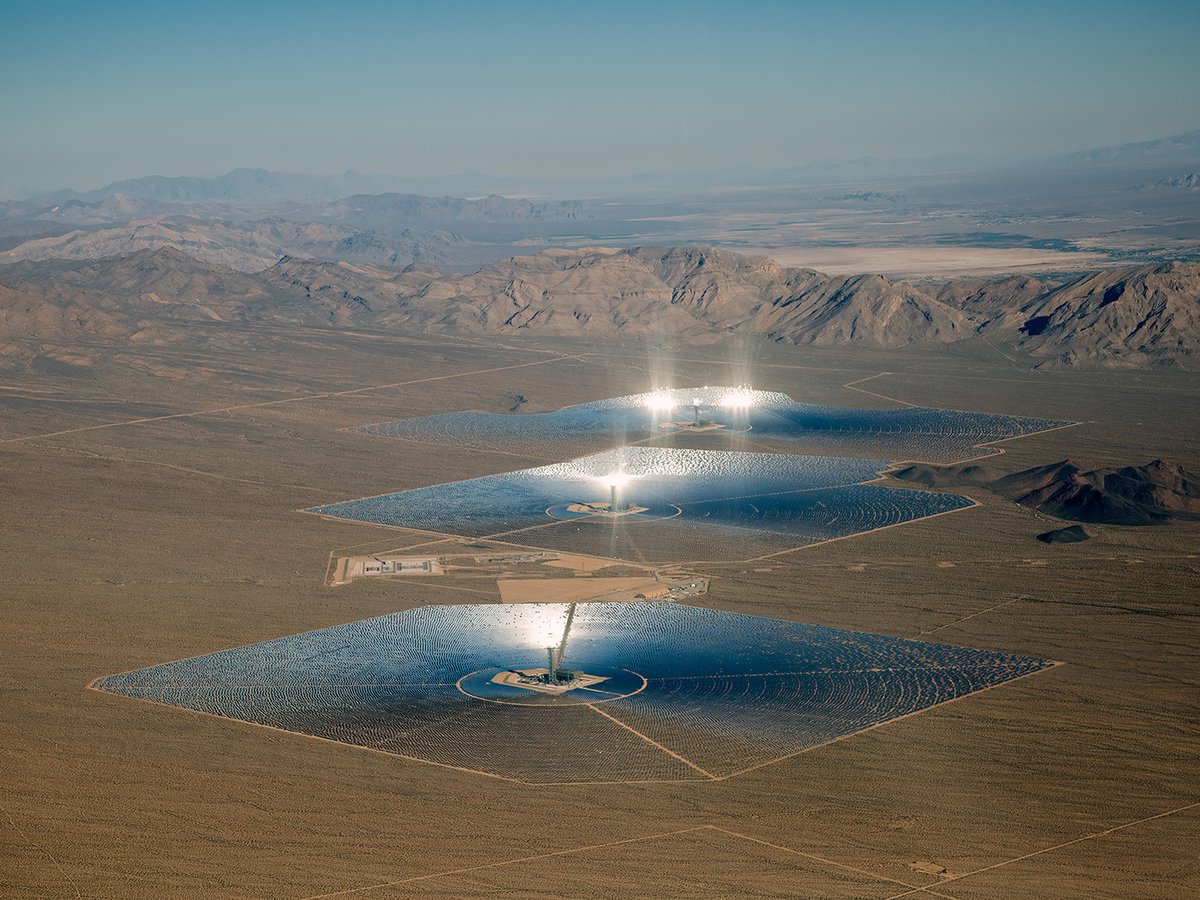 Aerial view of the Ivanpah Solar Electric Generating Station in the Mojave Desert, showing three power towers surrounded by large fields of heliostats reflecting bright sunlight onto the towers.