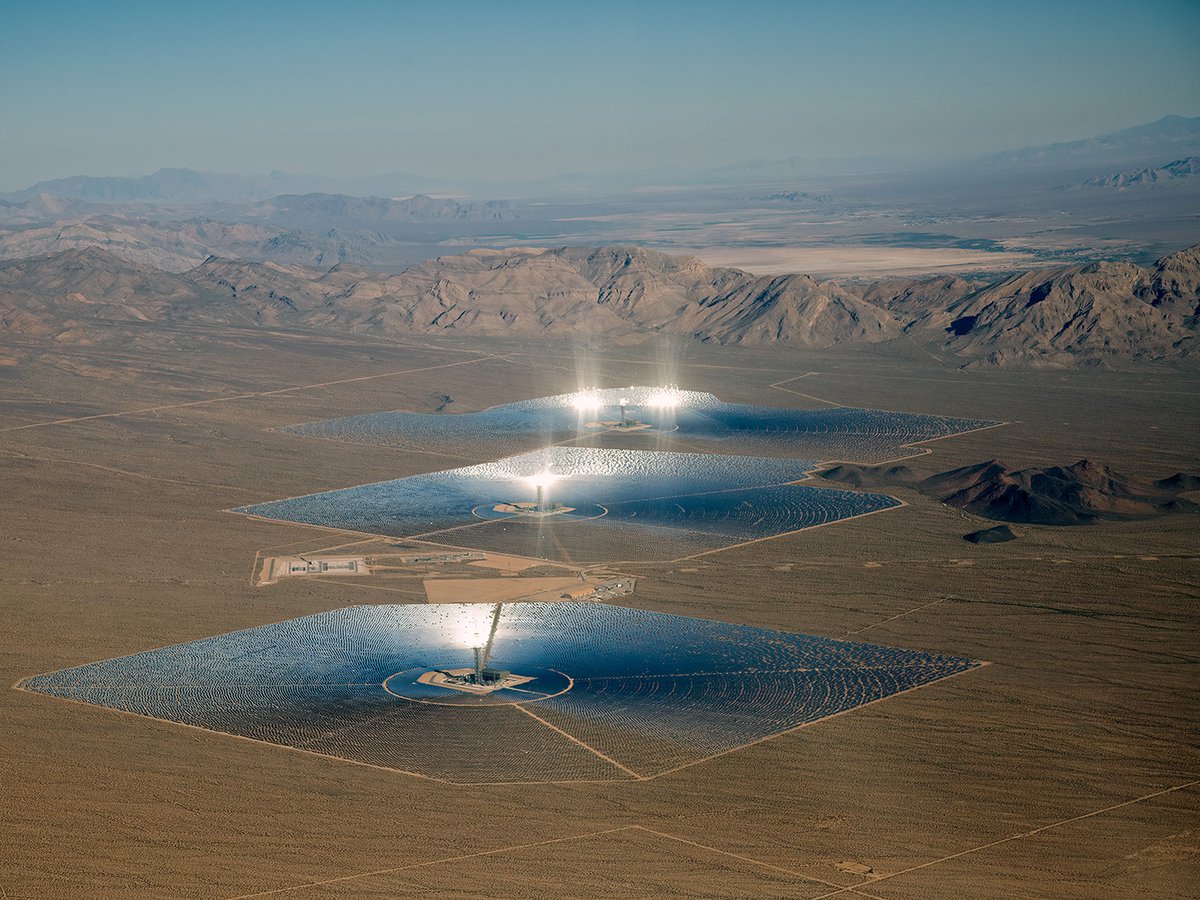 Aerial view of the Ivanpah Solar Electric Generating Station in the Mojave Desert, showing three power towers surrounded by large fields of heliostats reflecting bright sunlight onto the towers.