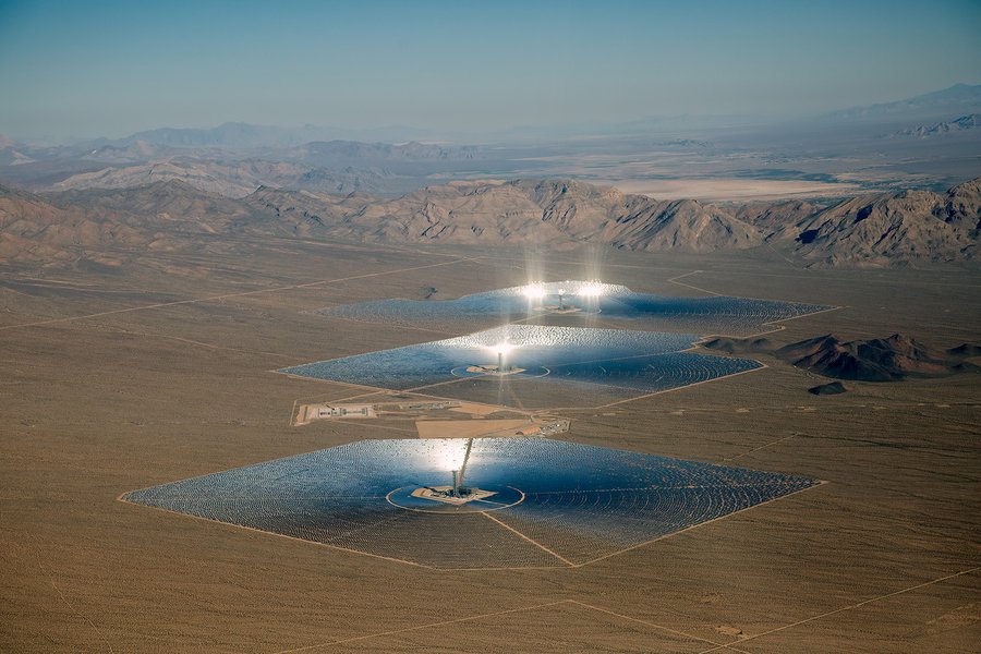 Aerial view of the Ivanpah Solar Electric Generating Station in the Mojave Desert, showing three power towers surrounded by large fields of heliostats reflecting bright sunlight onto the towers.