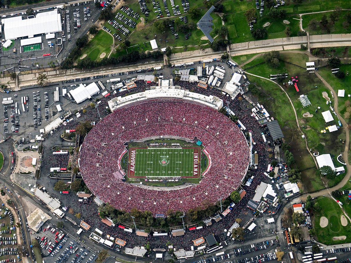 Aerial view of the Rose Bowl Stadium during the 2026 game, showing a full stadium with Indiana and Alabama logos in the endzones. A dark gray B-2 Spirit stealth bomber is captured in flight directly over the northern parking lot and Brookside Golf Club.