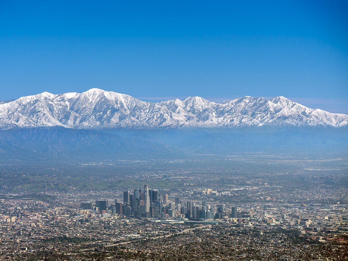 An aerial photo showing a breathtaking view of snow-capped mountains towering over the bustling metropolis of Downtown Los Angeles, marking a rare phenomenon during California's wet and cold winter season.