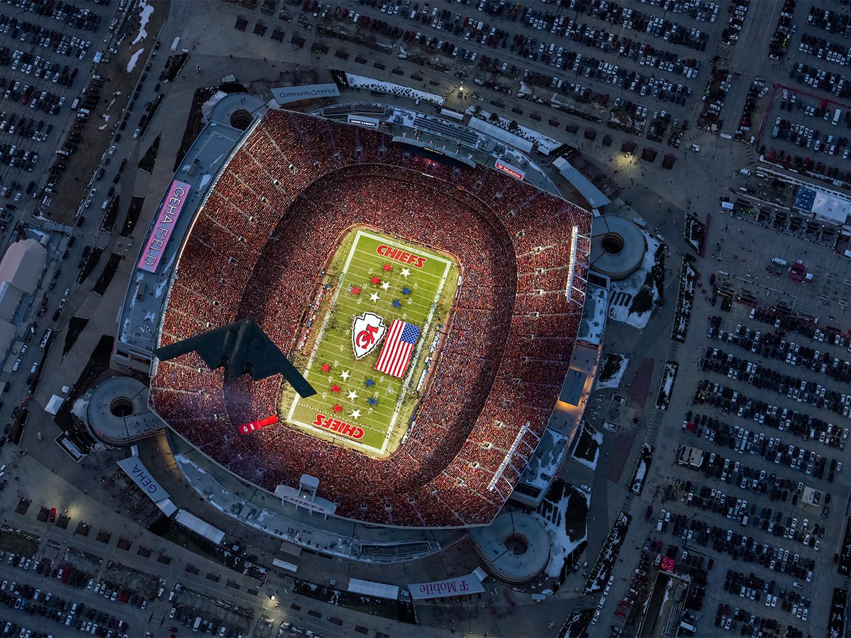An aerial photograph capturing a B-2 stealth bomber soaring over GEHA Field during the 2025 AFC Championship Game, with a vibrant sea of red Kansas City Chiefs fans filling the stands and a large American flag displayed on the illuminated field below.