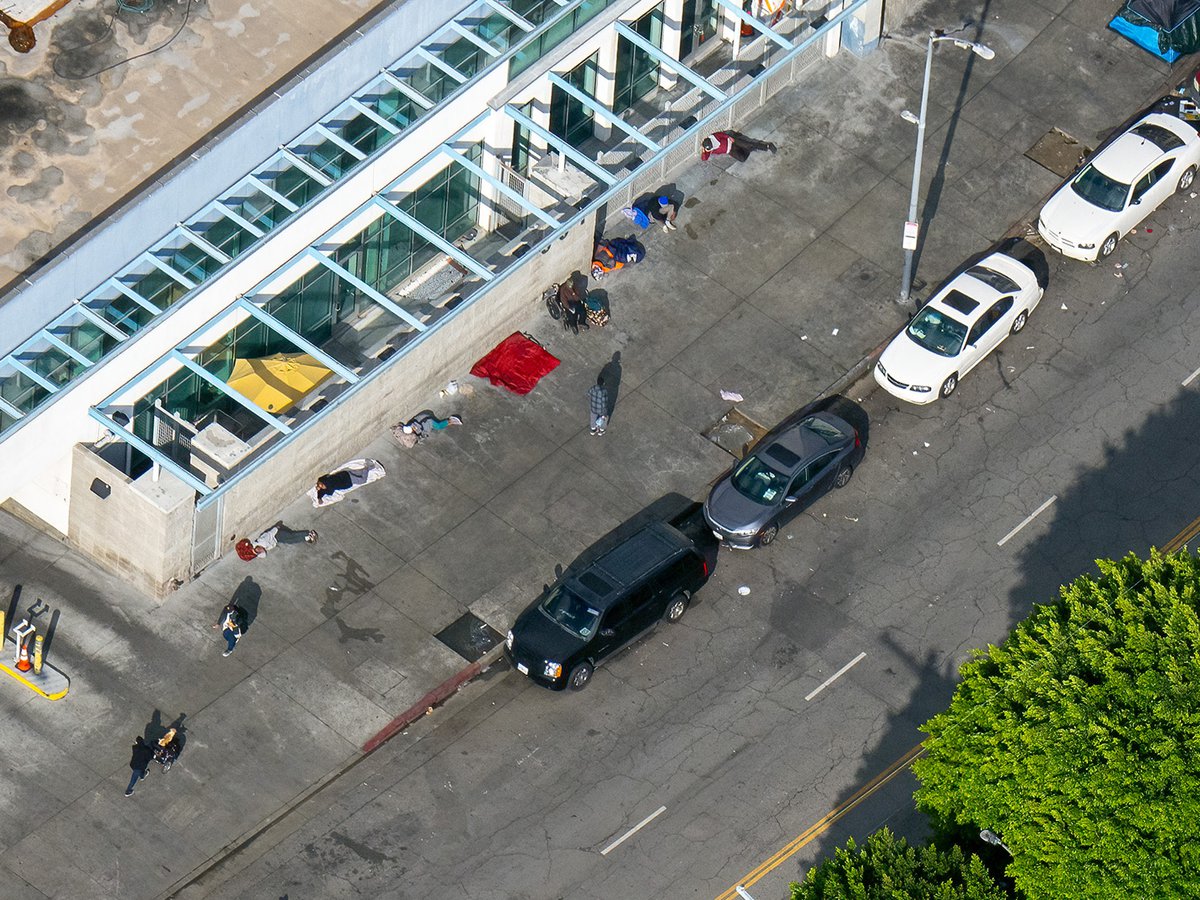 An aerial photo of Downtown Los Angeles shows a heart-breaking scene of numerous homeless people sleeping on blankets outdoors on the sidewalk, highlighting the unfortunate plight of those without safe, permanent shelter in the city.