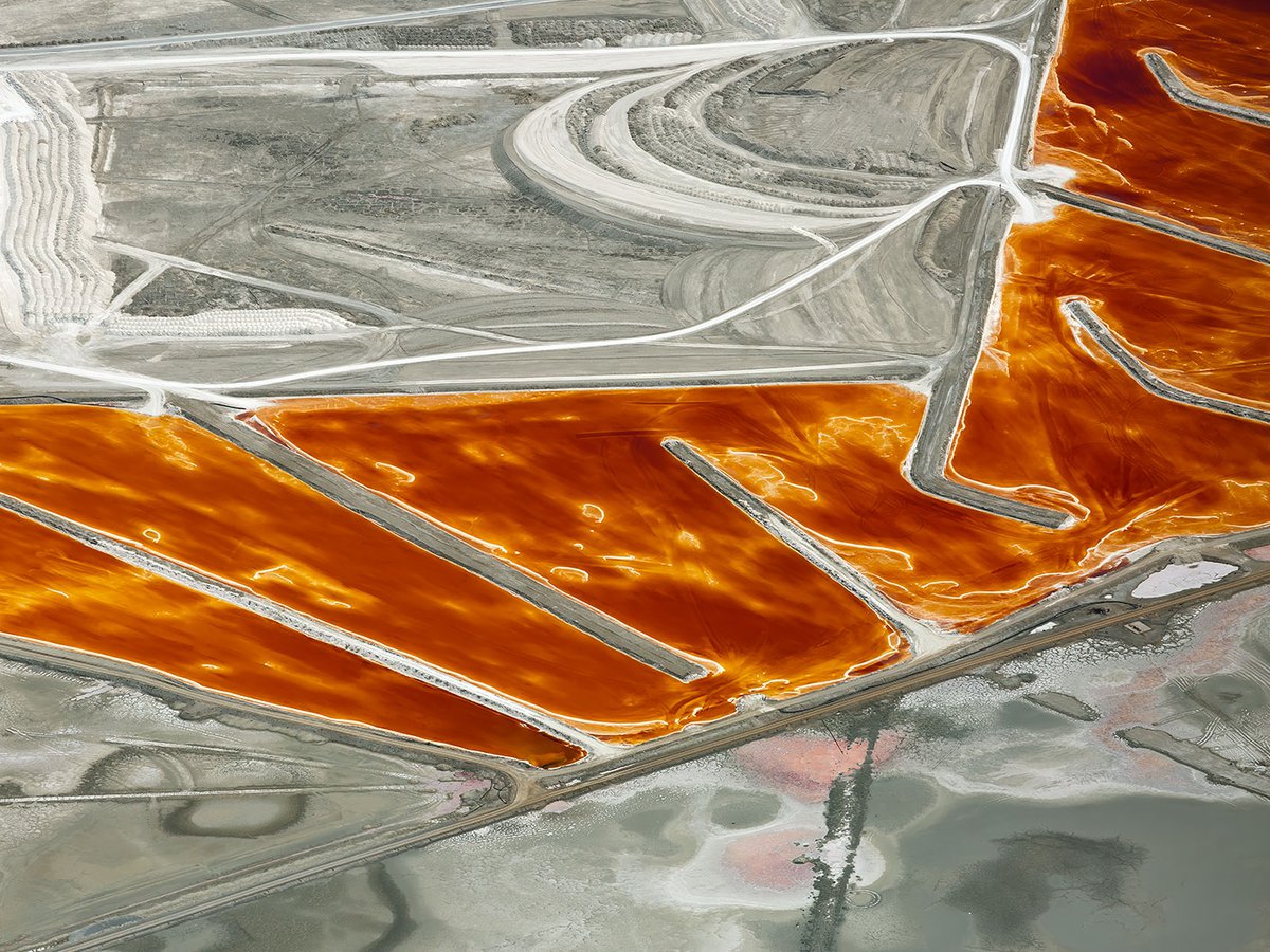 Aerial photo of the mineral pools of Searles Lake shimmering with fiery reds and oranges amidst a vast expanse of muted beige terrain, near Trona, California